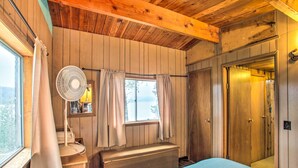 Un dormitori de cabana de fusta amb un llit, una banca, una finestra amb cortines, un ventilador i una porta.