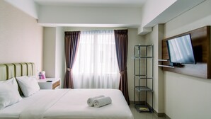 A hotel room with a bed, a TV mounted on the wall, a bedside table, and a window with curtains. - Comfort Studio Apartment At Padina Soho Residence (Tangerang)