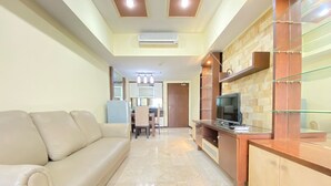 A modern living room with a beige sofa, a TV on a wooden cabinet, and a kitchen area in the background. - Spacious And Modern 2Br At Braga City Walk Apartment (Bandung)