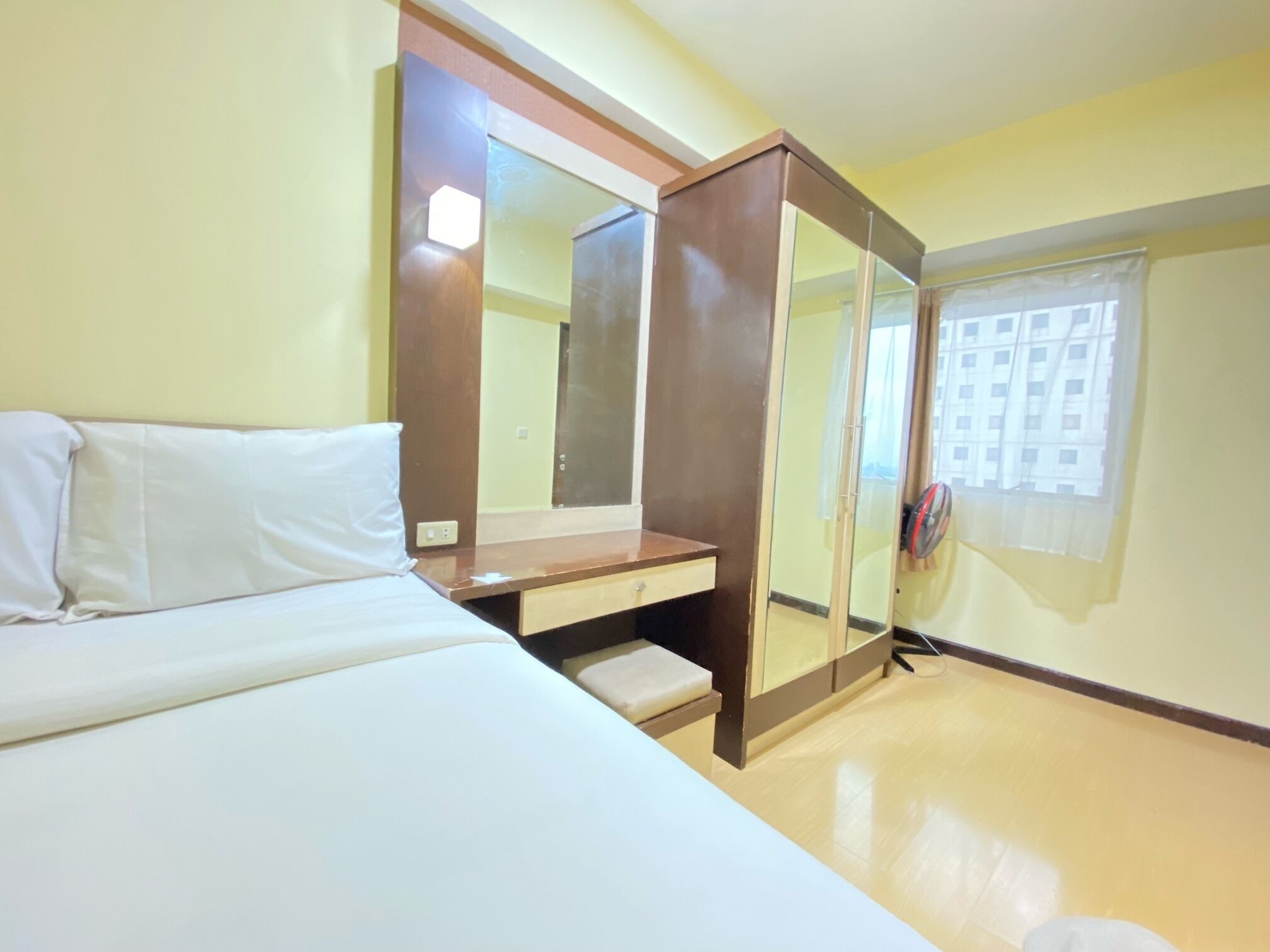 Habitación de hotel con cama, mesita de noche de madera, espejo y ventana con vista a edificios.