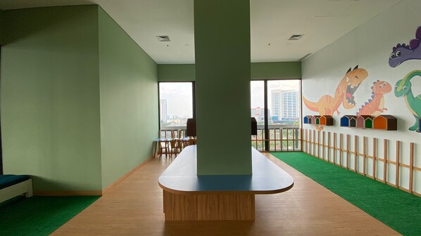 Una habitación con una mesa larga, sillas, un ventanal con vistas a edificios y una pared decorada con motivos de dinosaurios de colores.