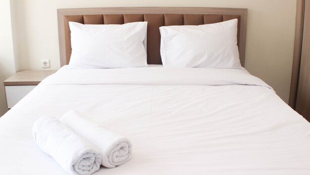Un letto rifatto con lenzuola e cuscini bianchi, un asciugamano bianco arrotolato su un lato e una testiera in legno.