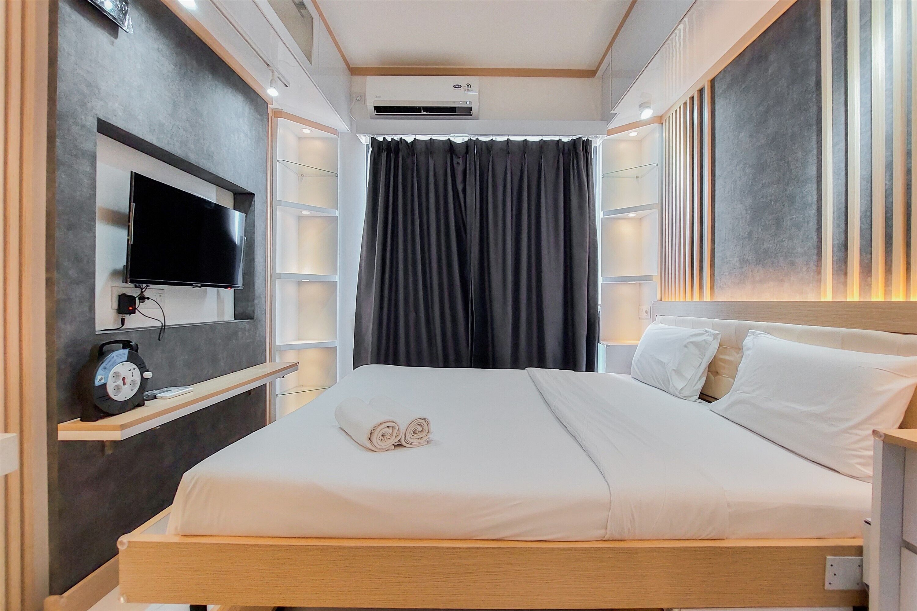 A modern hotel room with a flat-screen TV mounted on the wall, a bed with white linens, a bedside table with a clock, and a large window with dark curtains.