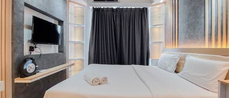 A modern hotel room with a flat-screen TV mounted on the wall, a bed with white linens, a bedside table with a clock, and a large window with dark curtains.