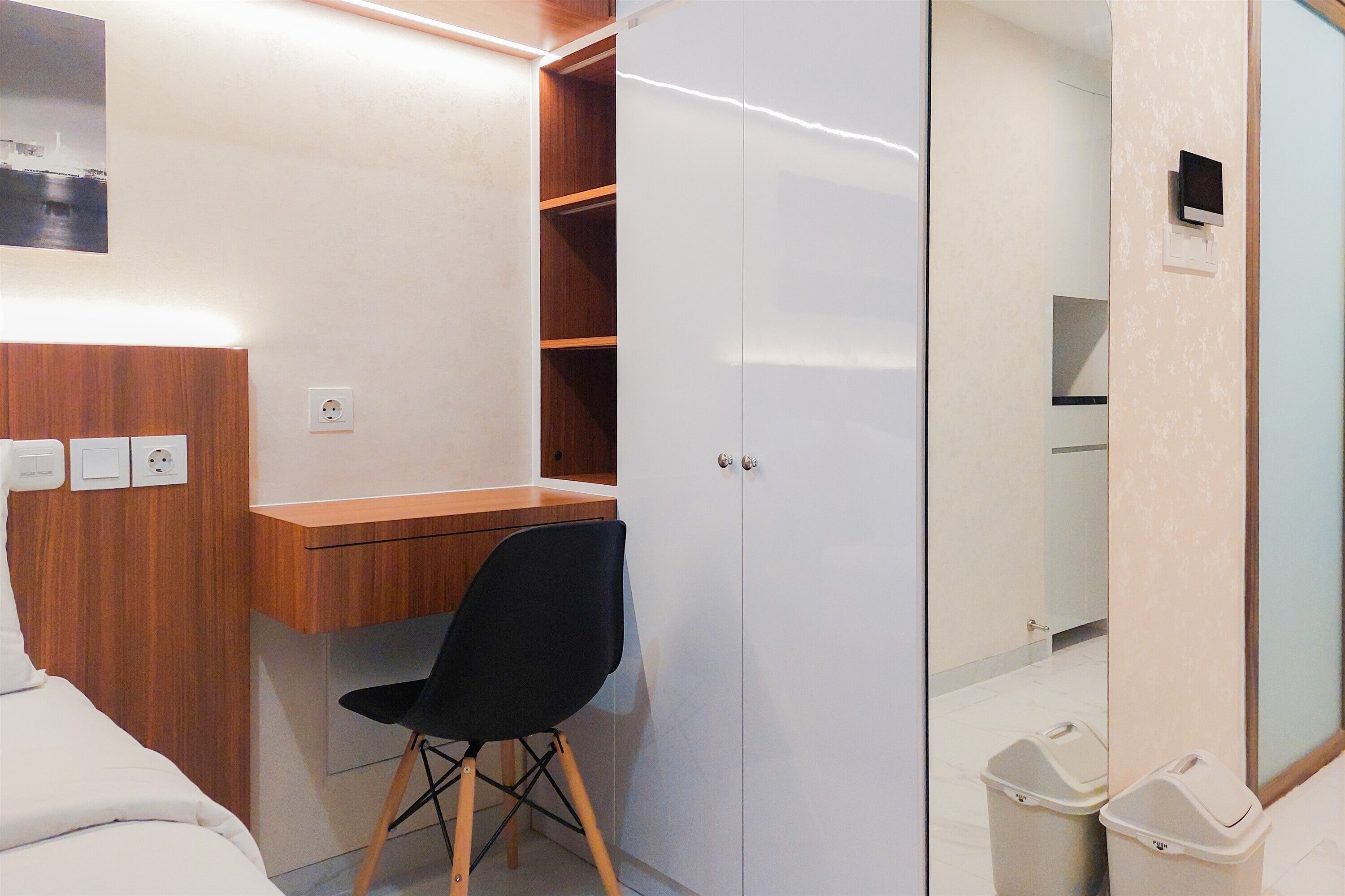 A bedroom with a wooden desk, a black chair, a wardrobe, and a small trash bin.