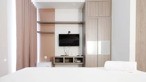 A modern hotel room with a bed, a TV mounted on the wall, a wooden wardrobe, and a small desk with a lamp. - Cozy Living Studio At Taman Melati Surabaya Apartment (Surabaya)