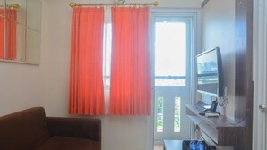 A room with a brown sofa, a television on a stand, a tray table, and a window with orange curtains. - Comfy And Best Deal 2Br At Green Pramuka City Apartment (Jakarta)