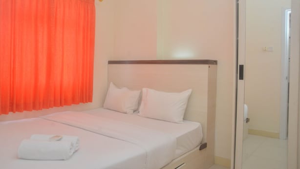 A hotel room with a bed, white bedding, an orange curtain, and a door to the right. - Comfy And Best Deal 2Br At Green Pramuka City Apartment (Jakarta)