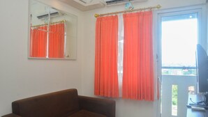 A room with a brown sofa, a large mirror, orange curtains, and a window with a view. - Comfy And Best Deal 2Br At Green Pramuka City Apartment (Jakarta)