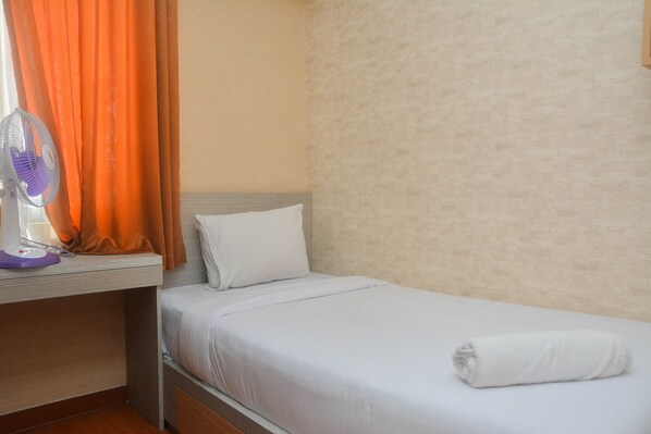 A single bed with white bedding, a white pillow, and a rolled white towel. There is a purple fan on a table next to the bed.