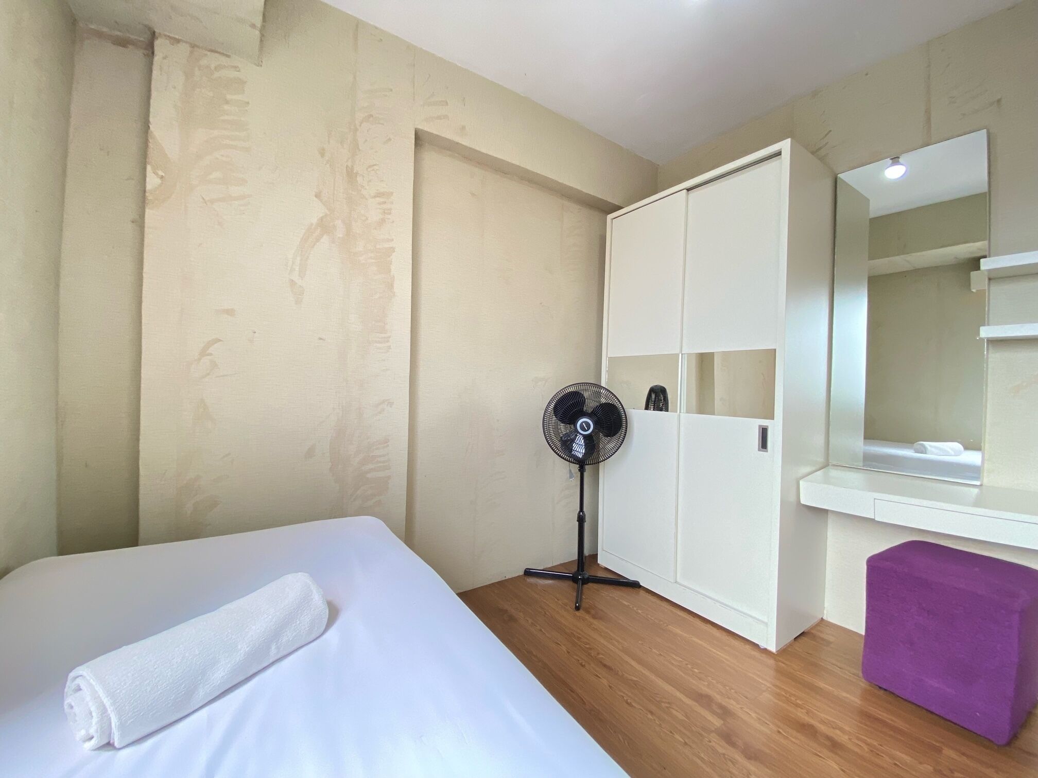 Une chambre d’hôtel avec un lit, un ventilateur sur pied, une armoire, un pouf violet et un miroir.