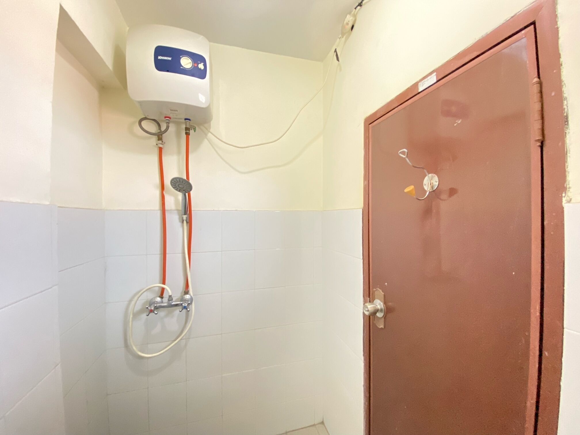A bathroom with a white and blue tiled wall, a white water heater, a shower with a hose, and a brown door.