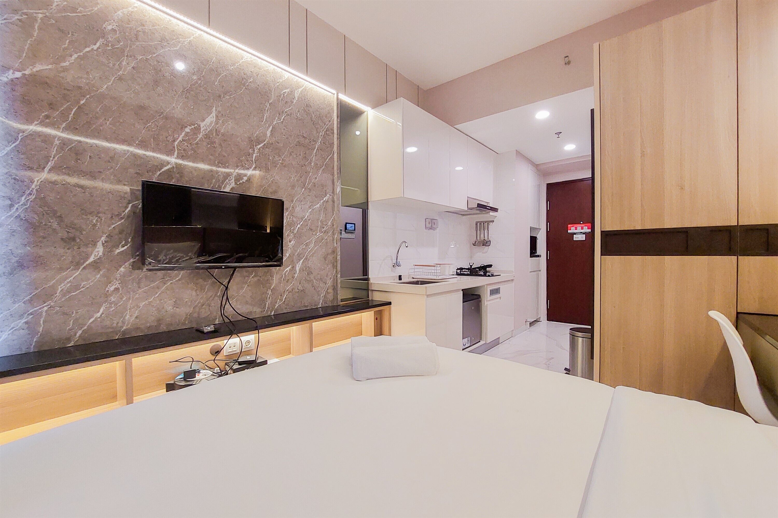 A modern hotel room with a marble wall, a flat-screen TV, a bed with a white pillow, a wooden wardrobe, and a kitchen area in the background.