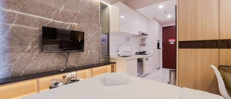 Una habitación de hotel moderna con pared de mármol, televisor de pantalla plana, cama con almohada blanca, armario de madera y una zona de cocina al fondo.
