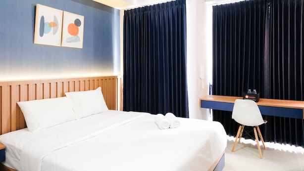 Habitación de hotel con cama, escritorio, silla y dos ventanas grandes con cortinas oscuras.