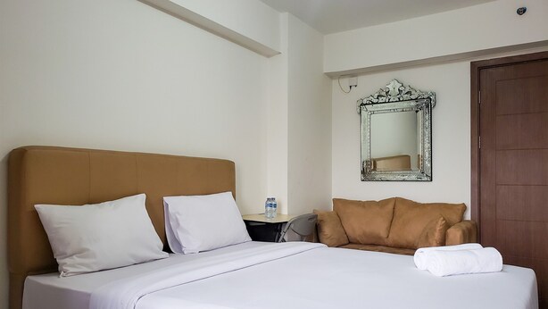 A hotel room with a bed, pillows, a nightstand, a water bottle, a mirror, and a door. - Warm And Cozy Studio At Cinere Resort Apartment (Depok)