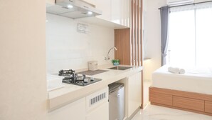 A modern kitchen with a stove, sink, and dishwasher, a small window with curtains, and a bed with a wooden headboard. - Comfy And Warm Studio Room At Sky House Bsd Apartment (Pagedangan)