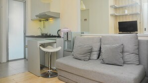 A modern kitchen with a built-in stove, a bar stool, and a grey sofa with cushions. - Minimalist And Warm 2Br At Bassura City Apartment (Jakarta)