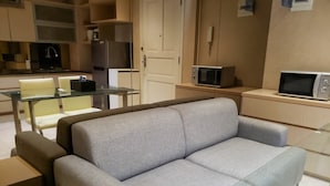 A modern living room with a grey sofa, glass coffee table, and a kitchen area with a fridge, microwave, and cabinets.