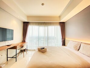 A hotel room with a bed, a desk, a television, and a view of the city through large windows. - Cozy Studio Room At Green Kosambi (Greko) Apartment (Bandung)