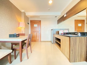 Quarto de hotel compacto com mesa de jantar, cadeiras, pequena área de cozinha com pia e bancada, e porta de madeira.