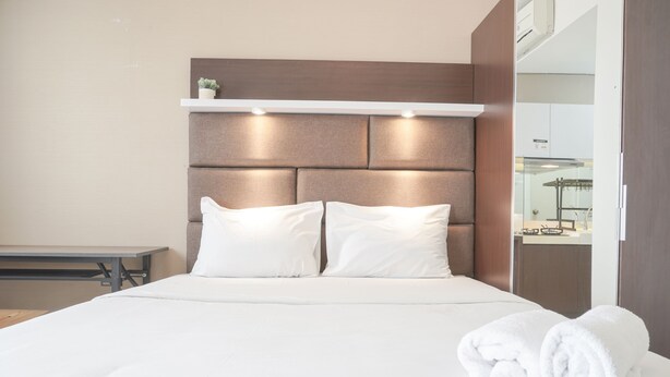 Un lit bien fait, avec du linge de lit blanc et des oreillers, une table de chevet et une étagère fixée au mur, sur laquelle se trouve une petite plante.