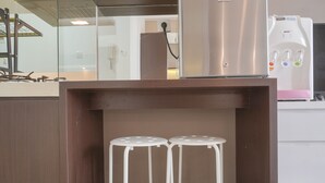 Un coin cuisine comprenant un réfrigérateur en acier inoxydable, un distributeur d’eau blanc et deux tabourets blancs placés sous un comptoir en bois.