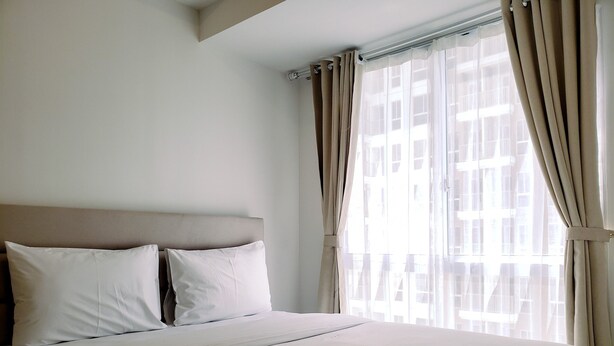 A hotel room with a large bed, two pillows, a headboard, and a window with curtains. - Minimalist And Tidy 2Br Apartment At Tokyo Riverside Pik 2 (Teluknaga)