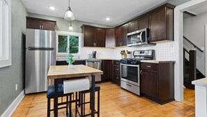 A modern kitchen with dark wood cabinets, stainless steel appliances, a wooden dining table with bar stools, and a view of the outdoors through a window.