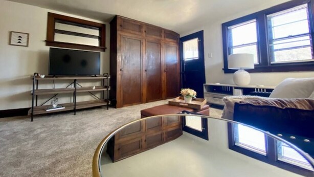 A living room with a TV, wooden cabinets, a sofa, and a window with blinds. - The Tiny Piney  - Live. Work. Play. Downtown Green Bay! (Green Bay)