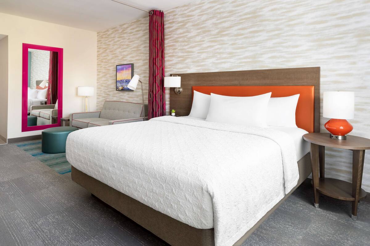 A hotel room with a large bed, a desk, a chair, a small table, and a mirror.