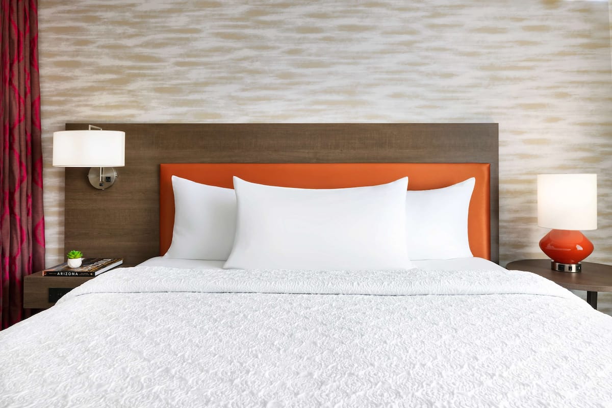 A hotel room with a bed, two nightstands, a red lamp, a small plant, and a book titled "ARIZONA".
