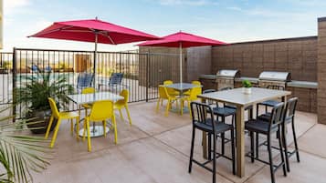 Een terras buiten met gele en zwarte stoelen, rode parasols en een barbecue.