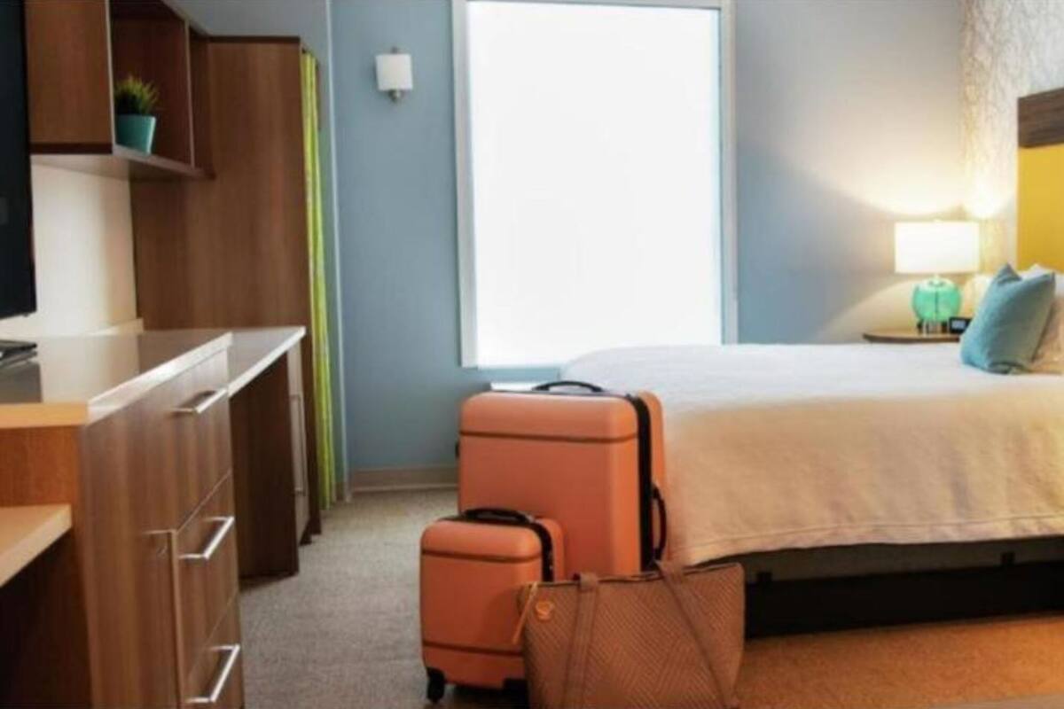 A hotel room with a bed, a desk, a chair, a suitcase, and a handbag.