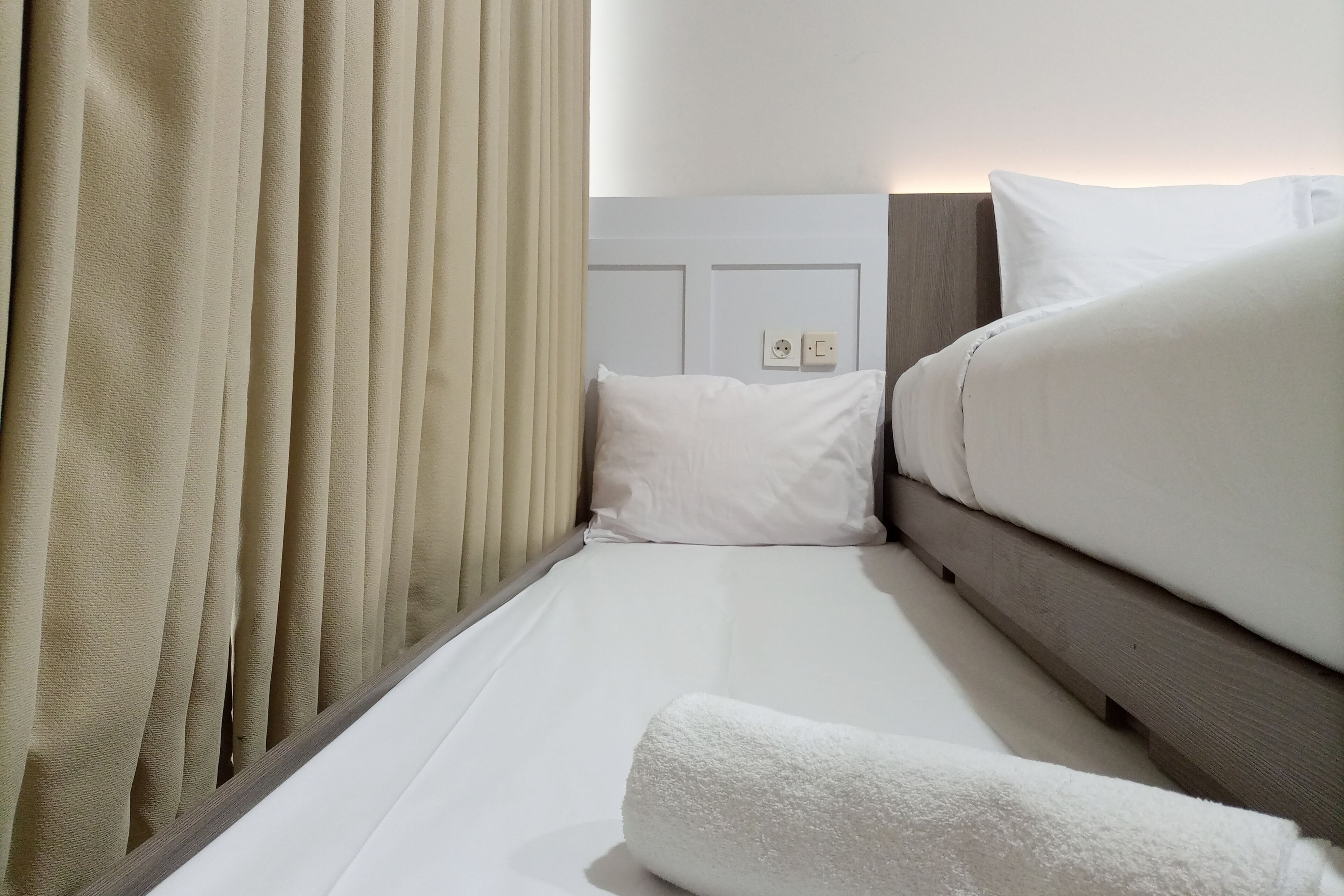 Een net opgemaakt bed met witte beddengoed en een opgerolde witte handdoek, een houten hoofdbord en een lichtschakelaar aan de muur.