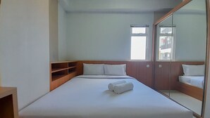Un lit bien fait, avec du linge de lit blanc et deux serviettes enroulées, une tête de lit en bois, un grand miroir et une fenêtre laissant entrer la lumière naturelle.