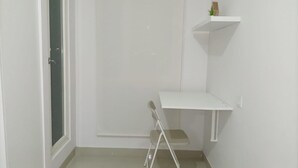 Une petite pièce blanche, avec un bureau, une chaise, une fenêtre et une porte.