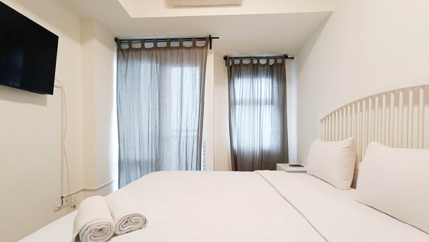A neatly made bed with a rolled towel, a bedside table, and curtains on the windows. - Cozy And High Floor Studio Room At Vida View Makassar Apartment (Makassar)