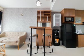 A modern living room with a sofa, two bar stools, a wooden coffee table, and a kitchen area with a refrigerator, microwave, and shelves.