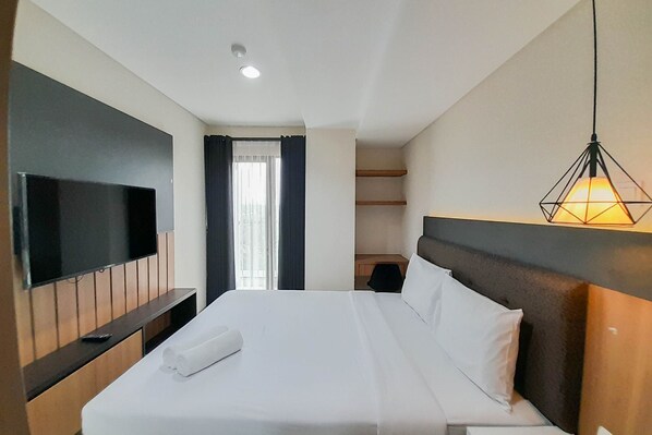 A hotel room with a large bed, a flat-screen TV mounted on the wall, a desk, and a hanging light fixture. - Fancy And Nice 1Br At Patraland Amarta Apartment (Ngaglik)