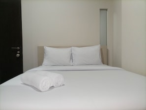 Una cama bien hecha con sábanas y almohadas blancas, una puerta oscura a la izquierda y una ventana pequeña a la derecha.