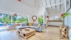 Ein großzügiger Wohnbereich mit einem großen, grauen Ecksofa, einem Holztisch und Blick auf einen Pool und Palmen.