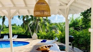 Ein Holztisch mit Bänken unter einer weißen Pergola, mit Blick auf einen Pool und üppiges Grün.