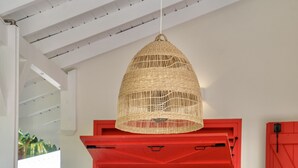 Ein Essbereich mit einem Holztisch, einer gewebten Pendelleuchte und einem Küchenblick durch ein rot gefasstes Fenster.