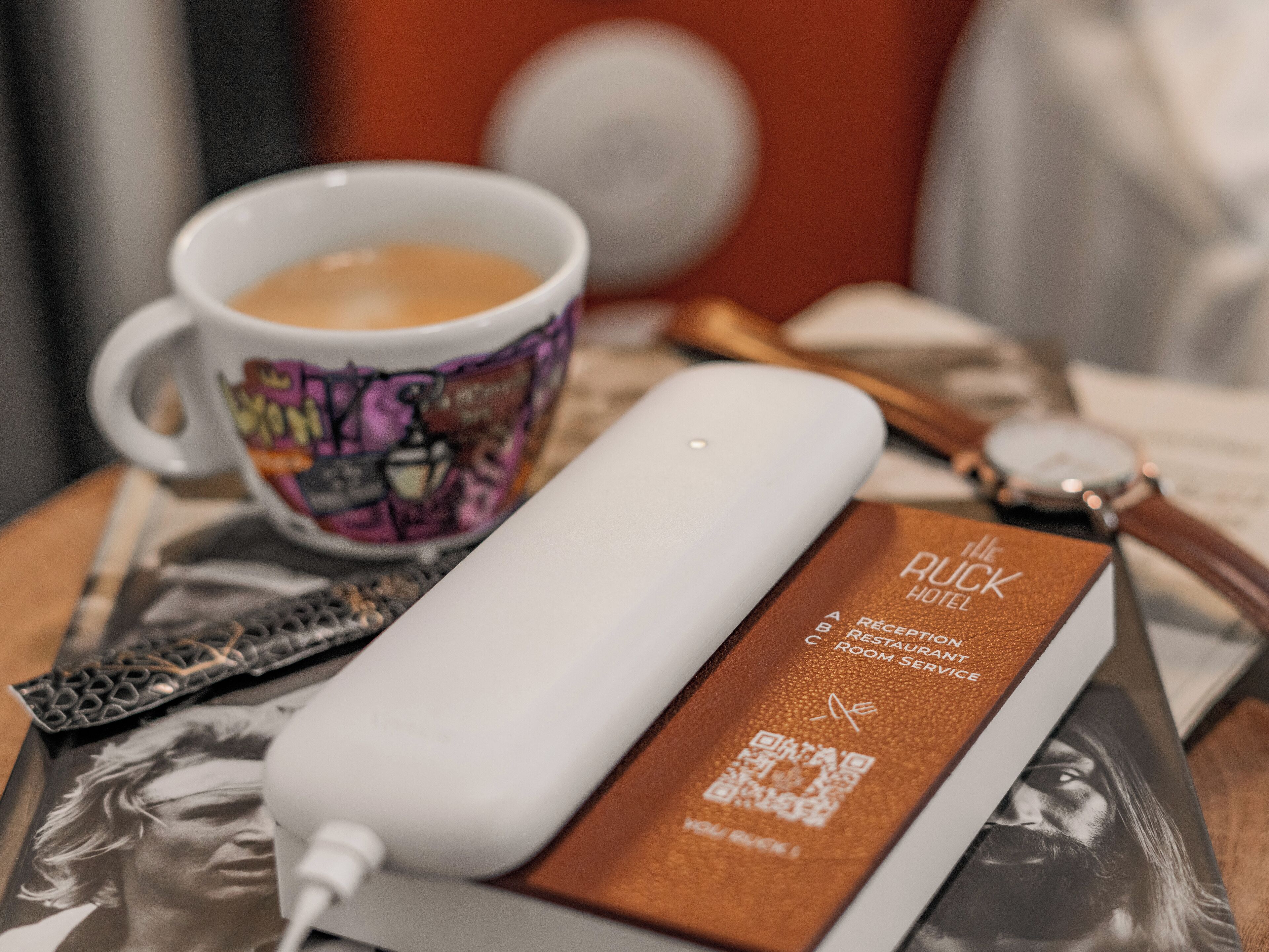 Una taza blanca con un diseño colorido, un cargador portátil blanco, un folleto marrón con código QR, un reloj y un bolígrafo sobre una mesa.