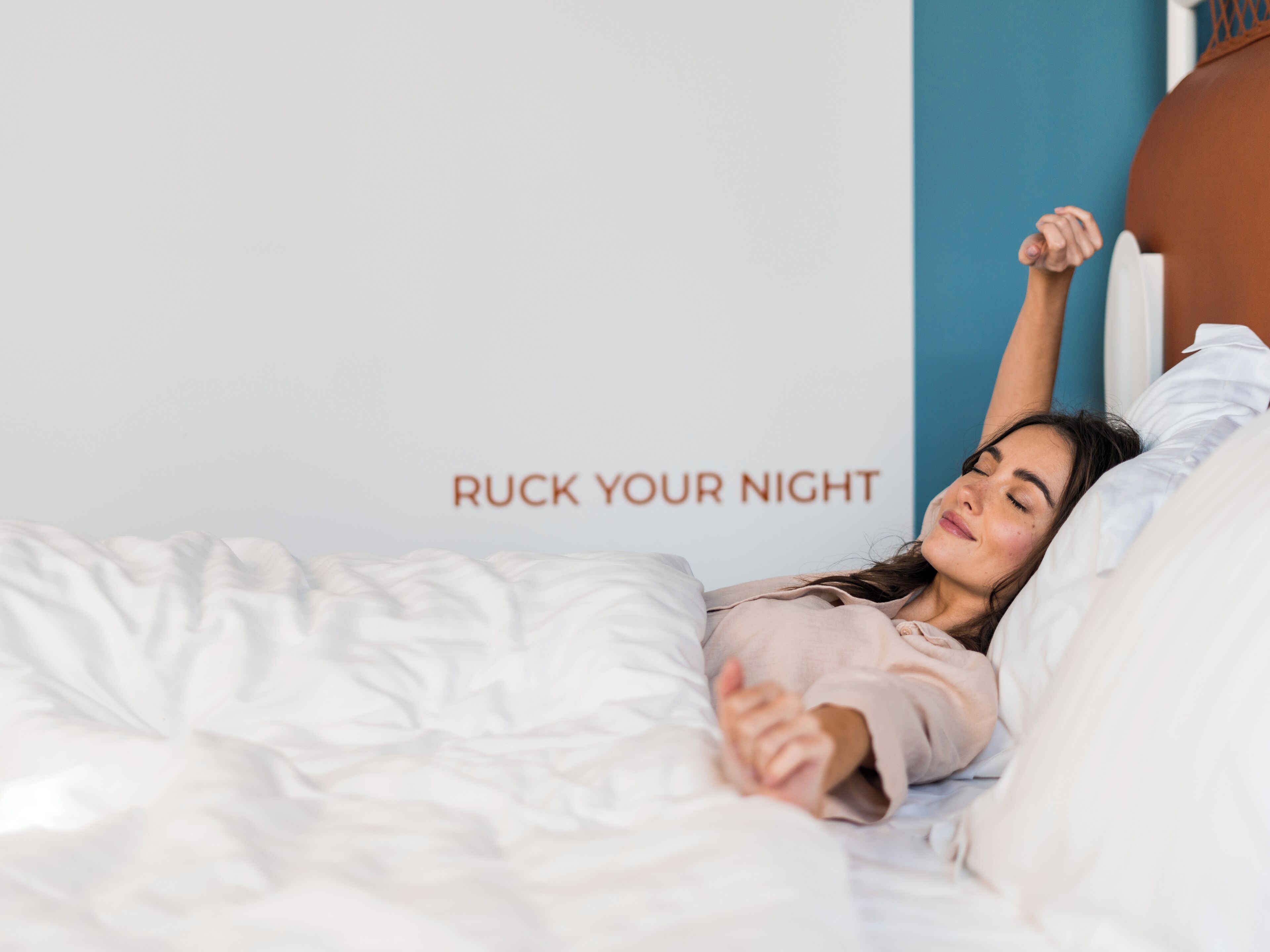 Een persoon ligt in bed met een kussen en een dekbed, en een muur met de tekst 'RUCK YOUR NIGHT'.