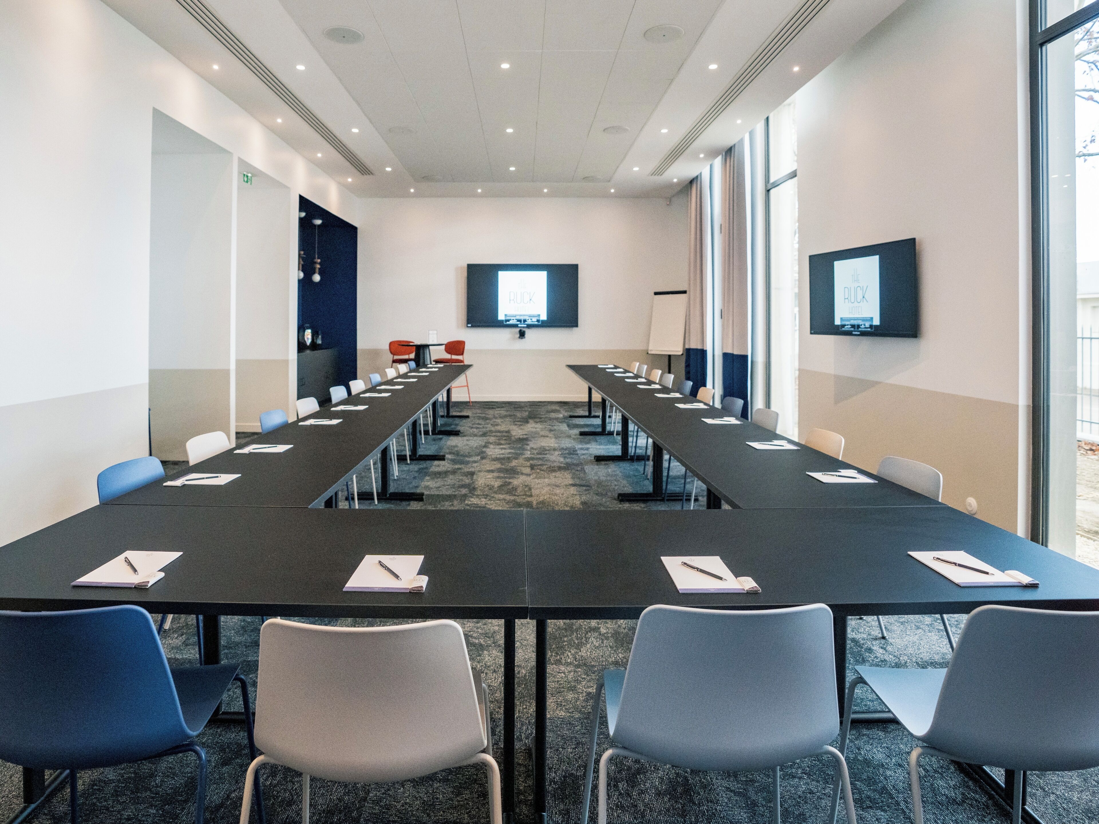 A conference room with a long table, chairs, a projector screen, and a television on the wall.