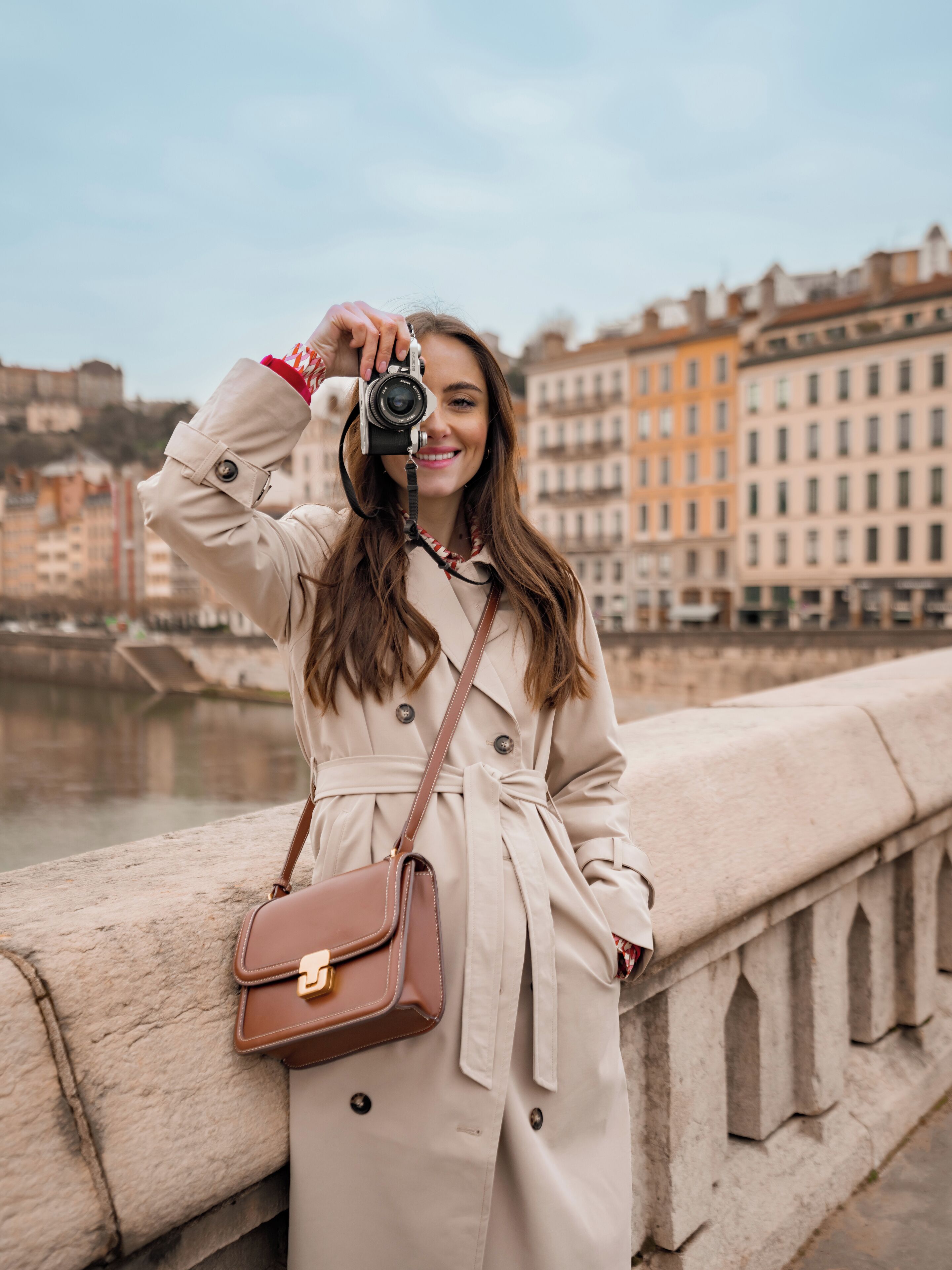 Une personne vêtue d’un trench-coat beige, tenant un appareil photo, se tient sur un pont avec des bâtiments en arrière-plan.
