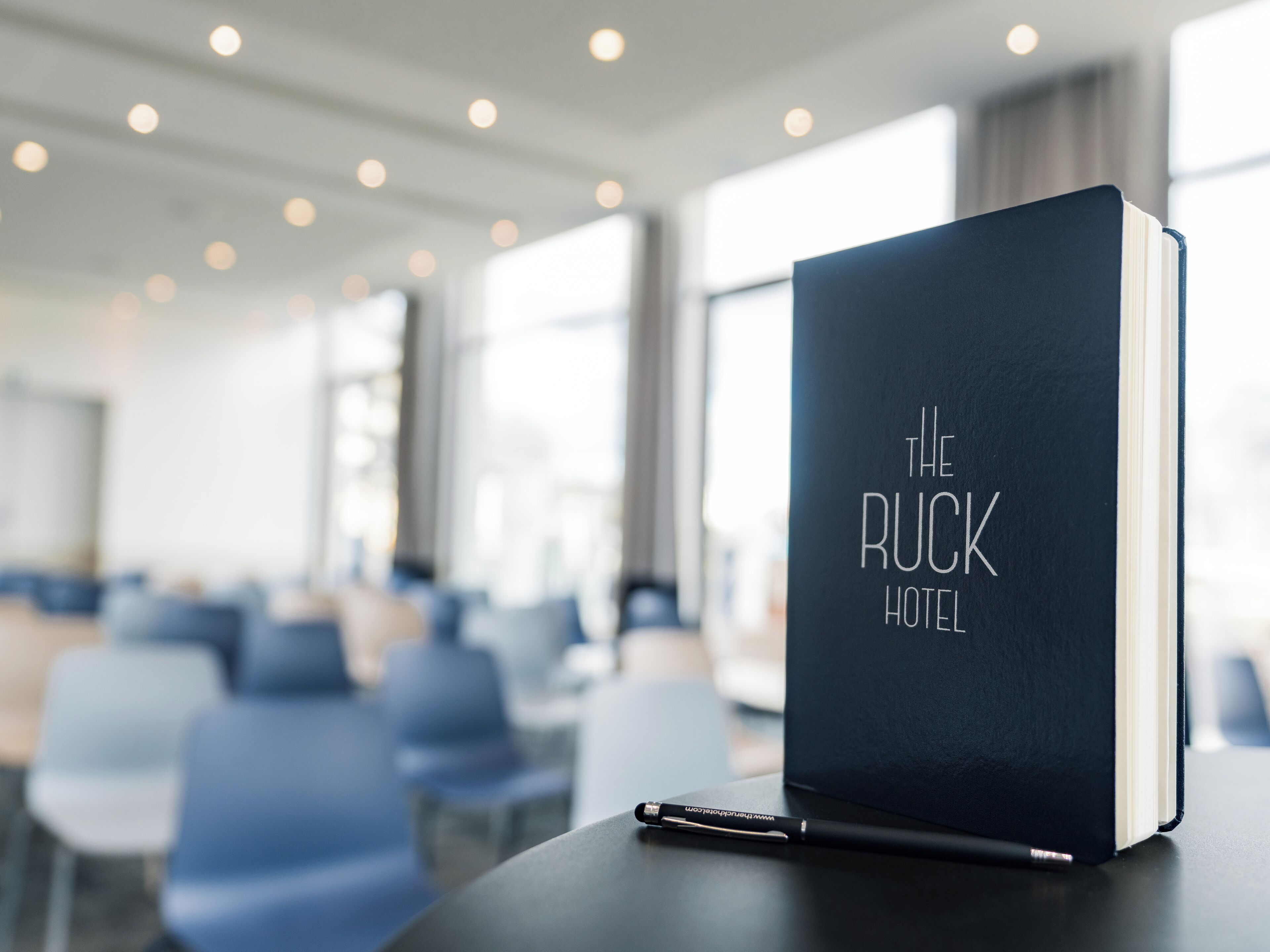 A black book titled "The Ruck Hotel" on a table in a hotel lobby with rows of chairs and large windows.