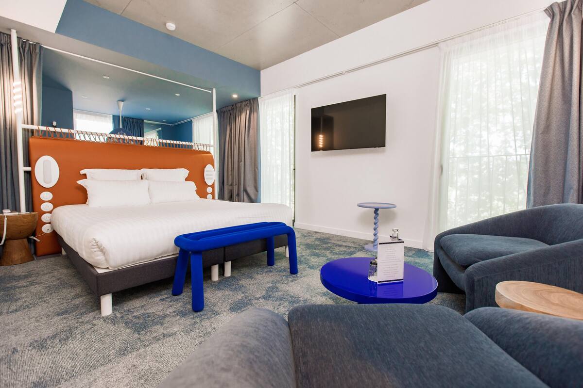 Een moderne hotelkamer met een groot bed, een blauwe bank, grijze banken, een televisie en een groot raam met gordijnen.